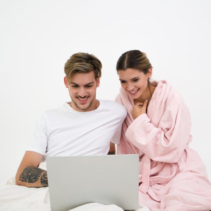 Man in White Crew-neck Shirt Sitting on Bed Beside Woman in Pink Bathrobe Looking at Laptop Computer